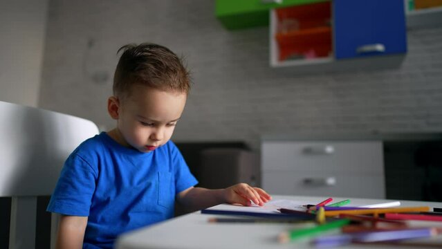 Caucasian toddler boy sits at the desk with lots of colorful pencils on. Little child picks one pencil and starts to draw. Low angle view.