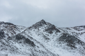 Snow-white alpine landscape with large snow-covered mountain range with sharp peaked top. Big snowy rocky ridge with pointy peak in cloudy sky. White snow on rocks. Pointed pinnacle of triangle shape.