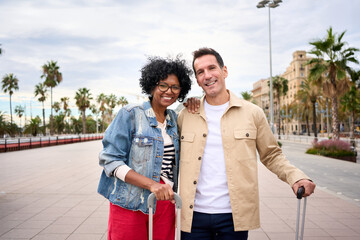 Portrait of cheerful middle-aged multiracial tourist couple embraced together look smiling at camera enjoying vacation tourism on city street. Caucasian husband and African wife for weekend getaway