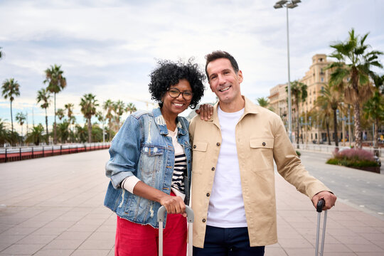Portrait of cheerful middle-aged multiracial tourist couple embraced together look smiling at camera enjoying vacation tourism on city street. Caucasian husband and African wife for weekend getaway