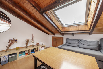 A small living room with sloping wooden ceilings with a large integrated skylight and light wooden...