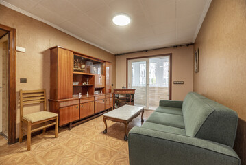 Small living room with antique shiny wooden furniture, green fabric sofa and terrace with aluminum enclosure in the background