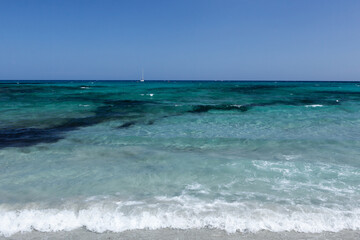 Clear blue water of the Mediterranean Sea with a white sailboat anchored on the horizon near the coast at Sardinia, Italy.
