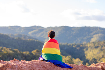 A young man sits on a mountain peak, wrapped in a rainbow flag, symbolizing LGBT pride month, with...
