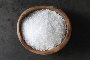 Natural salt in wooden bowl on dark grey table, top view