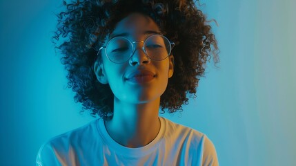 Attractive female student with curly hair wears transparent glasses white t shirt stands against blue background has calm face expression tender smile listens interlocutor with pleasur : Generative AI