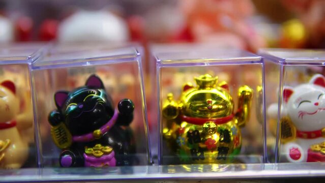 Variety of Maneki Neko cat statues places in transparent plastic cubes on the shop window filmed in closeup. Store in Vietnam with Traditional souvenirs moving one paw symbolizing luck and prosperity.