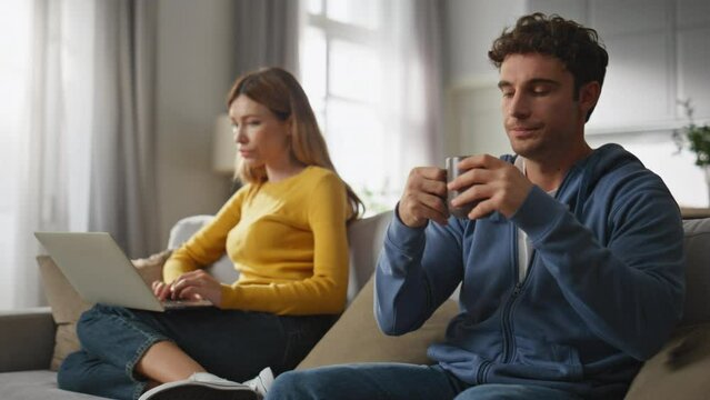 Offended couple ignoring each other at sofa. Bored man drinking coffee cup