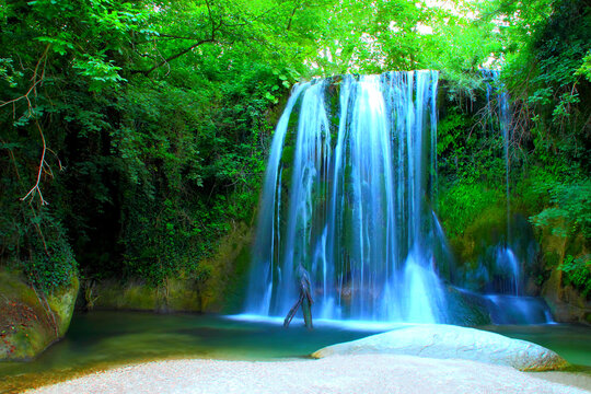 Long-exposure photo of Cascata "de lu Vagnat&ograve;" in Sarnano with glowing ethereal waters streaming downwards and collecting into an emerald basin below, between large rocks, the gravel shore and plants