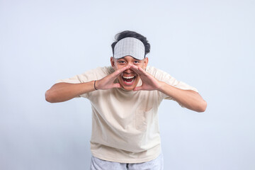 Young emotional man in sleep clothes and sleeping mask posing with shouting gesture against light...