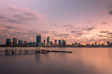 Jetty at South Perth foreshore, Western Australia overlooking city skyline. Water reflections on swan river with cloudy sky, pink, purple, blue hues mirrored on still water, buildings, city, dawn