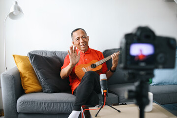 Senior Asian old man saying hi while playing guitar during podcast or live video broadcast,...