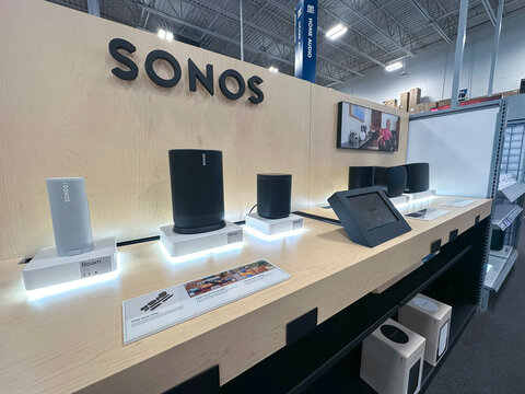 A variety of modern SONOS sound systems neatly displayed on a wooden shelf at an electronics retail store.