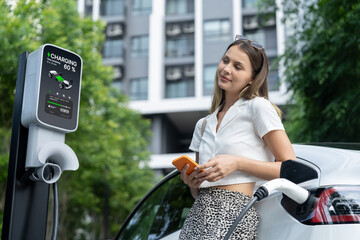 Young woman travel with EV electric car charging in green sustainable city outdoor garden in...