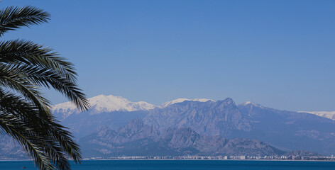 Turkey, Antalya presents a stunning view of the beautiful blue Mediterranean Sea, with towering snow-capped mountains in the distance and lush palm trees in the foreground.