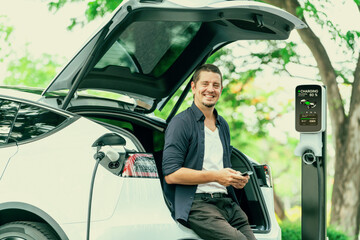 Man using smartphone online banking application to pay for electric car battery charging from EV charging station during vacation holiday road trip at national park or summer forest. Exalt