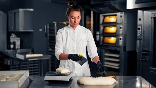 A female baker in uniform weighs evenly shaped dough on kitchen surface before baking in the oven