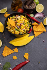 Freshly made mango salsa in a black ceramic bowl, nacho chips and ingredients
