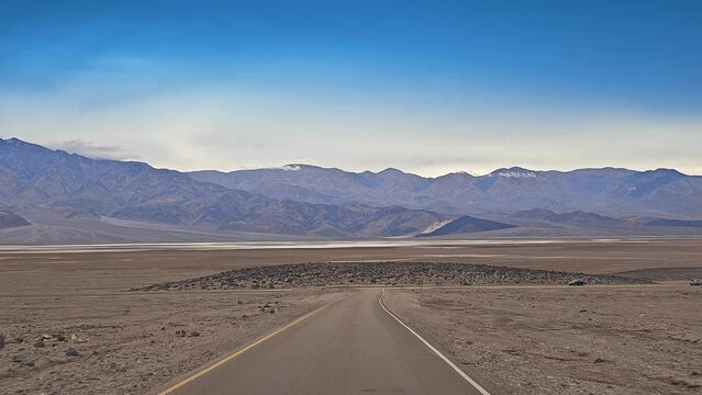 Drive Plate-POV-Death Valley-Approaching Badwater road from Artist Drive. Coming up on "T" intersection with shallow water of Lake Manly and mountains in distance under blue sky.