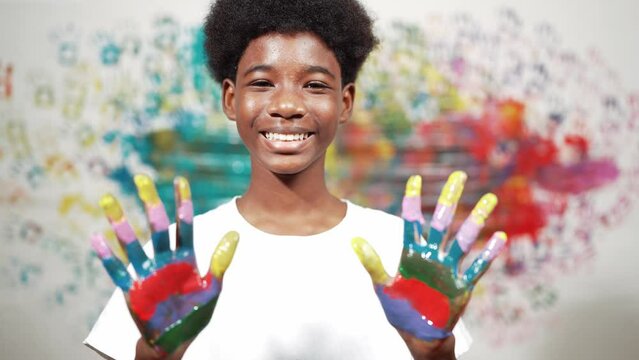 African boy looking at camera while showing two side of hand to camera with colorful color. Smiling happy high school teenager wearing white shirt while standing at colorful stained wall. Edification