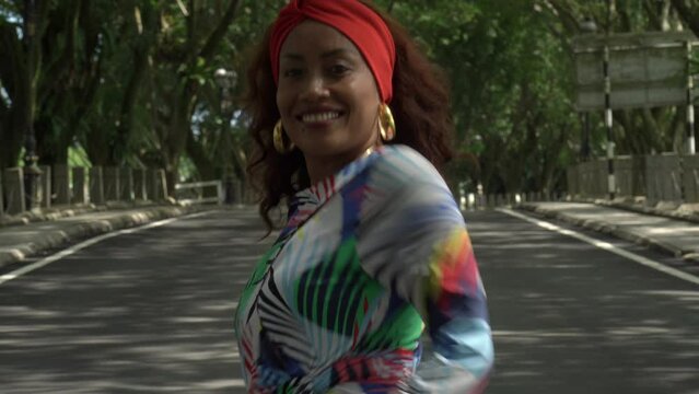 Asian woman in a colored stylish dress with turban walks and dance along the asphalt road under shady trees in the park. Portrait of a woman in motion between trees