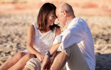 Fototapeta premium Portrait of romantic senior couple sitting on the beach at sunset enjoying vacation and retirement, two elderly people exchange cuddles expressing love and tenderness
