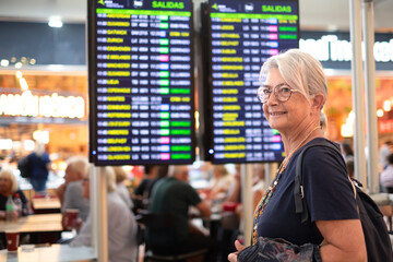 Woman traveler - senior lady standing inside airport terminal looking at timetable schedule. Travel and transportation themed image.