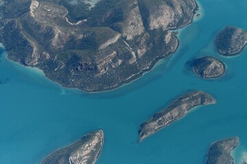 Uninhabited islands with rocky outcrops and covered in forest are surrounded by blue water. They are seen from the air, and the water colour changes wth the depth of the ocean.
