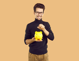 Young attractive smiling man in transparent glasses, dressed in black turtleneck and light trousers, throws coins into yellow piggy bank in the form of pig. Studio photo on isolated background.