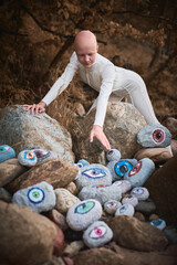 Young hairless girl with alopecia in white futuristic costume reaches hand for surreal landscape with lot of rocks with eyes, profound symbolism of embracing individuality in surreal world