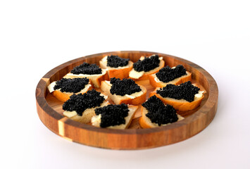 Black caviar on slices of bread lies on a round wooden plate on a white background
