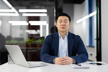 Serene Asian businessman practicing mindfulness meditation in modern office setting to alleviate stress and maintain focus.