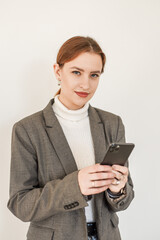 
a beautiful girl in a white turtleneck and a gray coat with red hair holds a phone in her hands