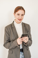 
a beautiful girl in a white turtleneck and a gray coat with red hair holds a phone in her hands