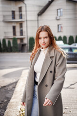 a beautiful woman with red hair in a gray coat walks through the city