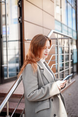 
a beautiful girl in a white turtleneck and a gray coat with red hair holds a phone in her hands