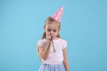 Birthday celebration. Cute little girl in party hat with blower on light blue background