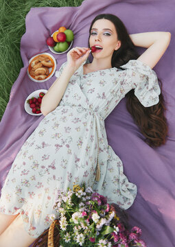 Picnic scene: pretty smiling girl lying on plaid in park and eating strawberry. Outdoor portrait of beautiful young woman in dress