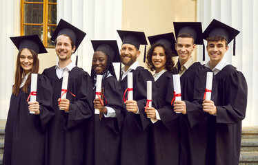 Group portrait of happy diverse college or university graduates. Seven multiracial male and female students in black grad caps and gowns standing outside their alma mater, holding diplomas and smiling