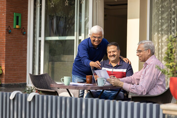 Elderly male friends smiling and reading paper and using laptop.