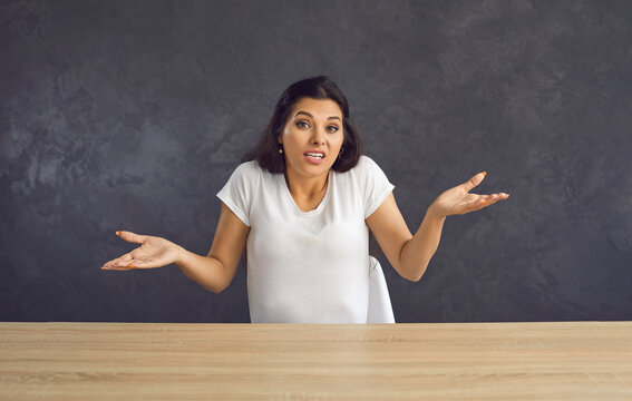 Young caucasian unsure woman sitting at a table shrugs saying that she can't help you. Beautiful brunette woman making a gesture to the camera which means I don't know. Grey studio background.