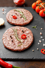Raw beef cutlets for burgers, rosemary, vegetables and spices on a stone plate on a dark background.