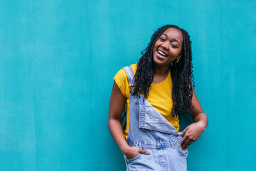 Cheerful young Afro-Latina with braids, wearing a vibrant yellow top and denim overalls, laughing...