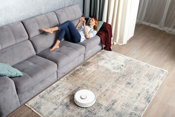 The girl lies on the sofa in the living room and controls a smart robot vacuum cleaner using a smartphone.