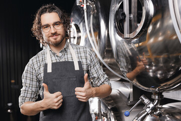 Portrait happy young bearded man shows thumbs up on background craft beer tank. Brewery worker with...