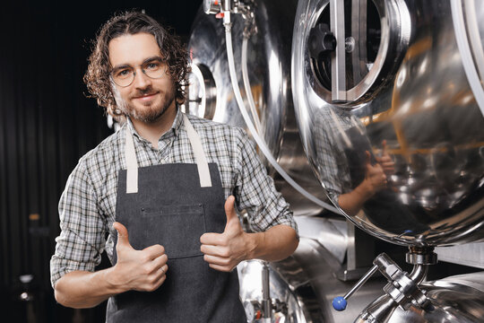 Portrait happy young bearded man shows thumbs up on background craft beer tank. Brewery worker with industrial equipment