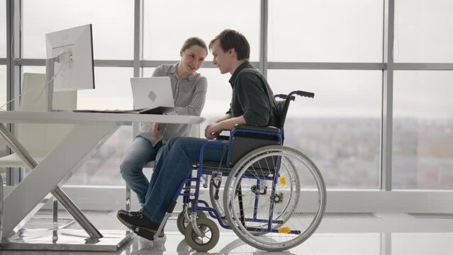 Paralyzed man work with woman in office. Colleagues discussing project together female shows to guy in wheelchair pie chart on paper document process. Workers discuss analytics data sit at desk.