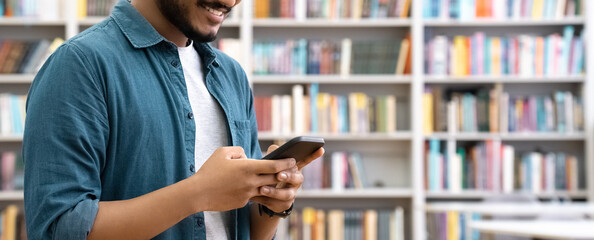 Banner of smiling Indian student chatting by mobile phone in university library. Young man using social media application app, browsing online on smartphone cellphone device gadget for studying. 