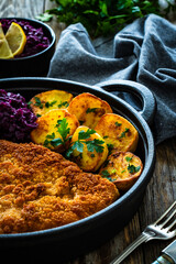Crispy breaded fried cutlet with baked potatoes and cooked red cabbage on wooden table
