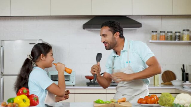 Joyful Indian father with kid dancing together at kitchen while cooking - concept of weekend holidays, family bonding and happy parenting